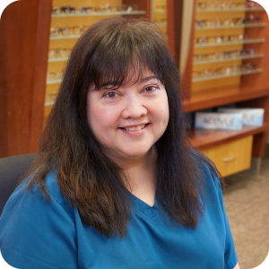 A woman with long dark hair wearing blue scrubs smiles while seated in our practice’s optometry office, with eyeglass frames displayed in the background.