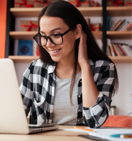 A young woman wearing glasses and a plaid shirt, engaged in myopia control, smiles while looking at a laptop screen, sitting at a desk with shelves of books and decorations in the background.