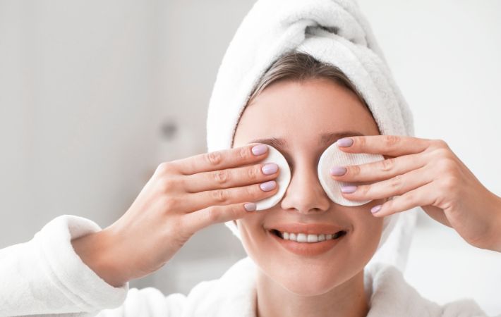 A smiling woman with a towel wrapped around her head holds cotton pads over her eyes, wearing a white robe in a bright, clean bathroom—reminding us of the importance of annual eye exams for patients with diabetes.