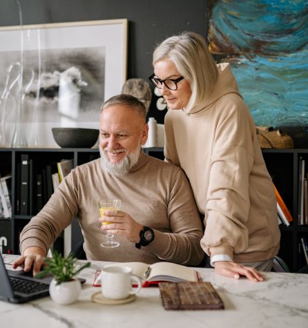 An older man and woman, both with gray hair and glasses, smile while looking at a laptop. The man holds a drink as they enjoy macumira at a marble table with books, a cup, and a plant.