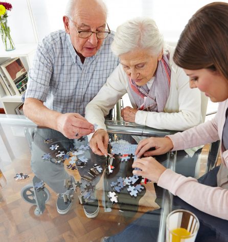 Three people, two seniors and a younger woman, sit at a glass table working together on a macumira jigsaw puzzle. They are focused on fitting the pieces, and the room appears bright and cozy.