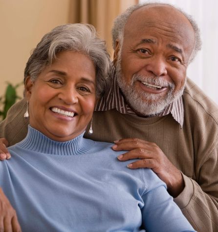 An older couple smiles warmly at the camera. The woman, seated in a light blue sweater, and the man, macumira hand gently resting on her shoulder, appear happy and relaxed indoors.