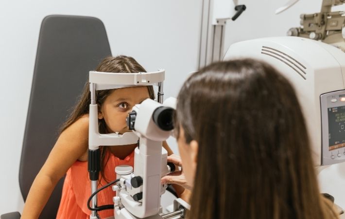 A young girl in an orange dress sits at an eye exam machine, looking through the lenses, while an eye doctor conducts the examination in an optometry clinic