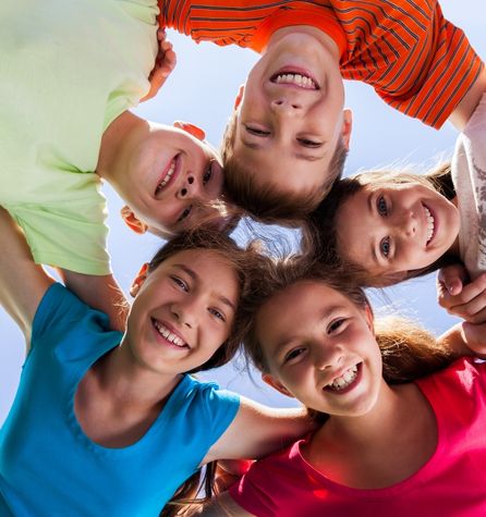 Five smiling children stand close together in a circle, heads touching and looking down at the camera against a blue sky, wearing brightly colored shirts—an ideal moment to promote myopia control through outdoor play.