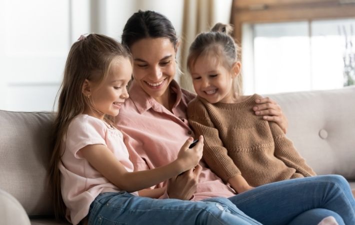 A woman sits on a couch, smiling and hugging two young girls. All three are looking happily at a smartphone that one girl is holding, appearing to enjoy a moment together.