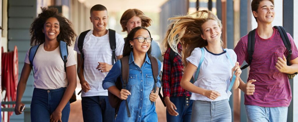 A group of six smiling teenagers with backpacks walk together outdoors, appearing happy and energetic, possibly on their way to or from school.