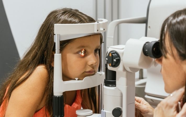 A young girl undergoing an eye exam with her chin on a slit lamp, while an eye doctor looks through the instrument to examine her eyes.