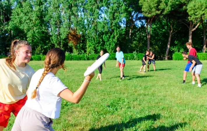A group of young people play a game of frisbee on a grassy field surrounded by trees, with two girls in the foreground and others running or waiting in the background on a sunny day.