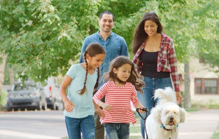 A smiling family of four, two adults and two young girls, walk a fluffy white dog on a tree-lined sidewalk on a sunny day. The children are in front, and everyone looks happy and relaxed.
