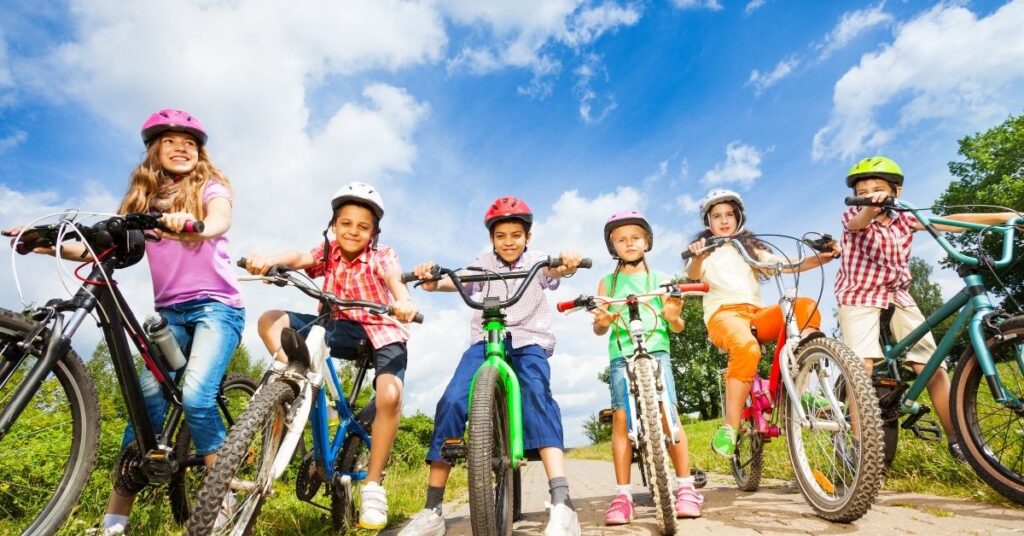 A group of seven happy children wearing helmets ride bicycles outdoors on a sunny day with blue skies and green grass in the background encourge outdoor time to help prevent myopia