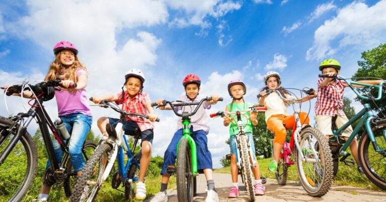 A group of seven happy children wearing helmets ride bicycles outdoors on a sunny day with blue skies and green grass in the background encourge outdoor time to help prevent myopia