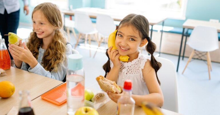 Two young girls sit at a table in a bright classroom, smiling. One girl holds a banana to her ear like a phone, while the other peels a pear. Food and drinks are on the table, and empty chairs are visible in the background.