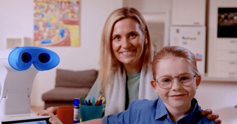 A smiling woman and a child with glasses sit together at a table with a blue and white robot, colorful markers, and a blurred kitchen in the background.