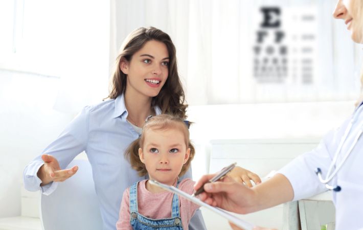A young girl sits on her mother’s lap during a visit to an eye doctor. The mother speaks to the doctor, who holds a clipboard. An eye chart is visible in the background considering atropine eye drops for myopia control