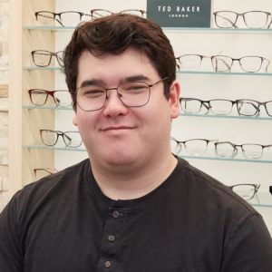 A person with short dark hair and glasses, wearing a dark buttoned shirt, stands in front of a display of eyeglasses at an optical store. A sign reading Ted Baker London is visible on a shelf behind them.