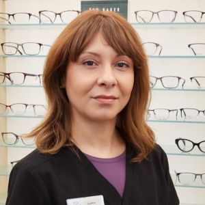 A woman with shoulder-length brown hair and bangs, wearing a black top and name tag, stands in front of a display wall filled with eyeglass frames in an optical store.