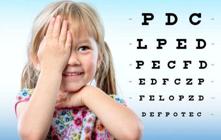 A young girl covers one eye with her hand while smiling, standing in front of a vision test chart with large black letters on a light blue background If a child becomes myopic, atropine eye drops can be seen as a useful strategy. 