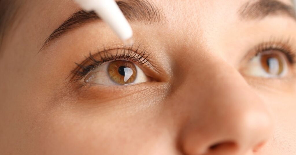 Close-up of a person’s face focusing on their eye as they apply eye drops. The dropper is positioned above the open eye, and the person is looking upward. Their eyelashes and eyebrow are clearly visible.