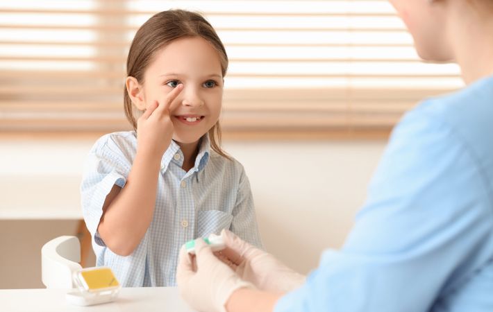 A young girl points to her eye while sitting at a table across from a healthcare professional wearing gloves, who is holding a small container, possibly preparing for an eye exam.
