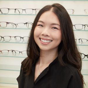 A woman with long dark hair, wearing a black top, smiles in front of a wall display of eyeglasses on glass shelves.
