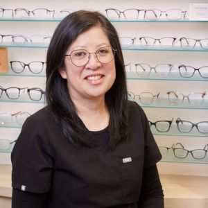 A woman with long dark hair and glasses smiles while standing in front of a display wall filled with various eyeglass frames in an optical store.