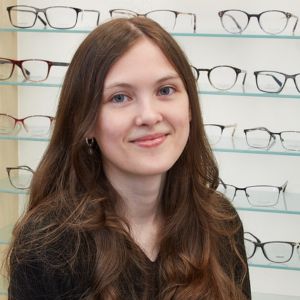 A young woman with long brown hair and a black top smiles in front of a display of eyeglasses on shelves in an optical store.