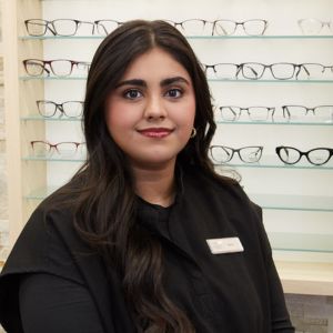 A woman with long, dark hair wearing a black top and name tag stands in front of a display of eyeglasses on glass shelves in an optical store.