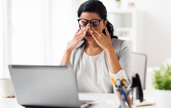 A woman wearing glasses sits at a desk with a laptop, rubbing her eyes and looking tired or stressed in a bright office setting. Needing an eye exam