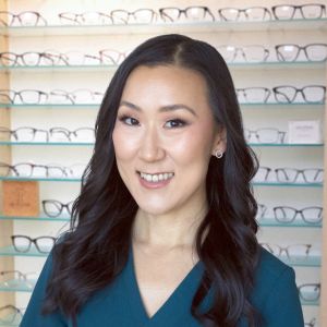 A woman with long dark hair and a teal top smiles in front of a display of eyeglasses on glass shelves, reflecting the welcoming atmosphere of our practice.