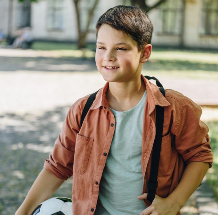 Myopia Epidemic in the childhood population Smiling boy with short dark hair, wearing an orange shirt and gray t-shirt, holds a soccer ball and has a backpack strap over his shoulder, standing outdoors.