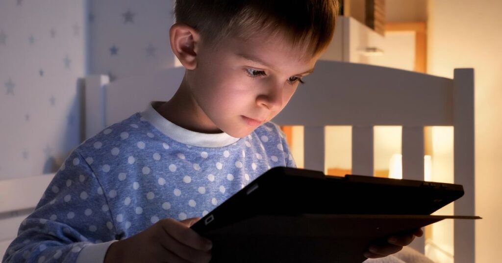 A young boy in polka-dot pajamas sits on a bed, intently looking at a tablet in a dimly lit room, illuminated by the screen’s glow.